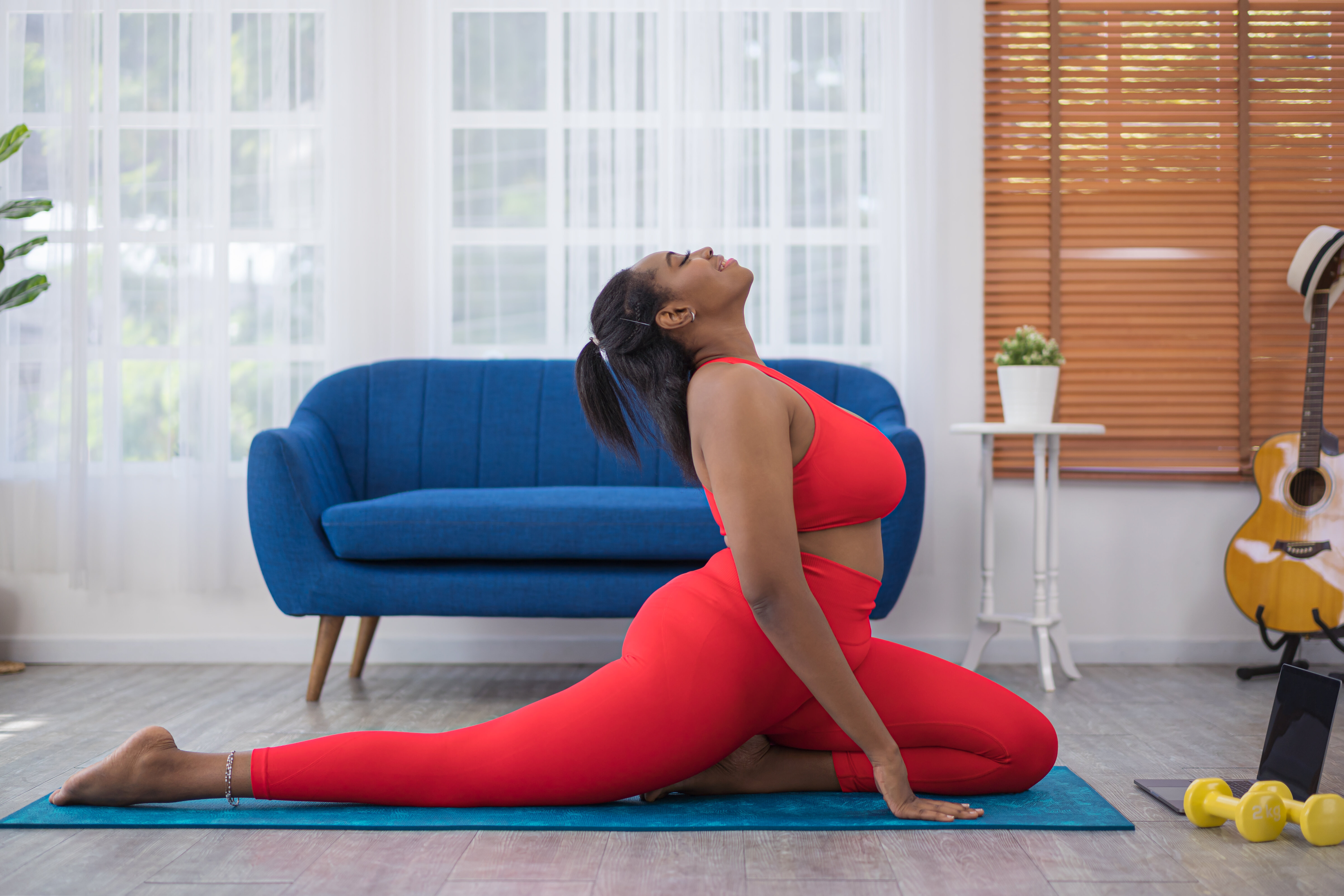 Woman in a red yoga outfit performing a pigeon pose in a living room with a blue couch, guitar, and dumbbells in the background