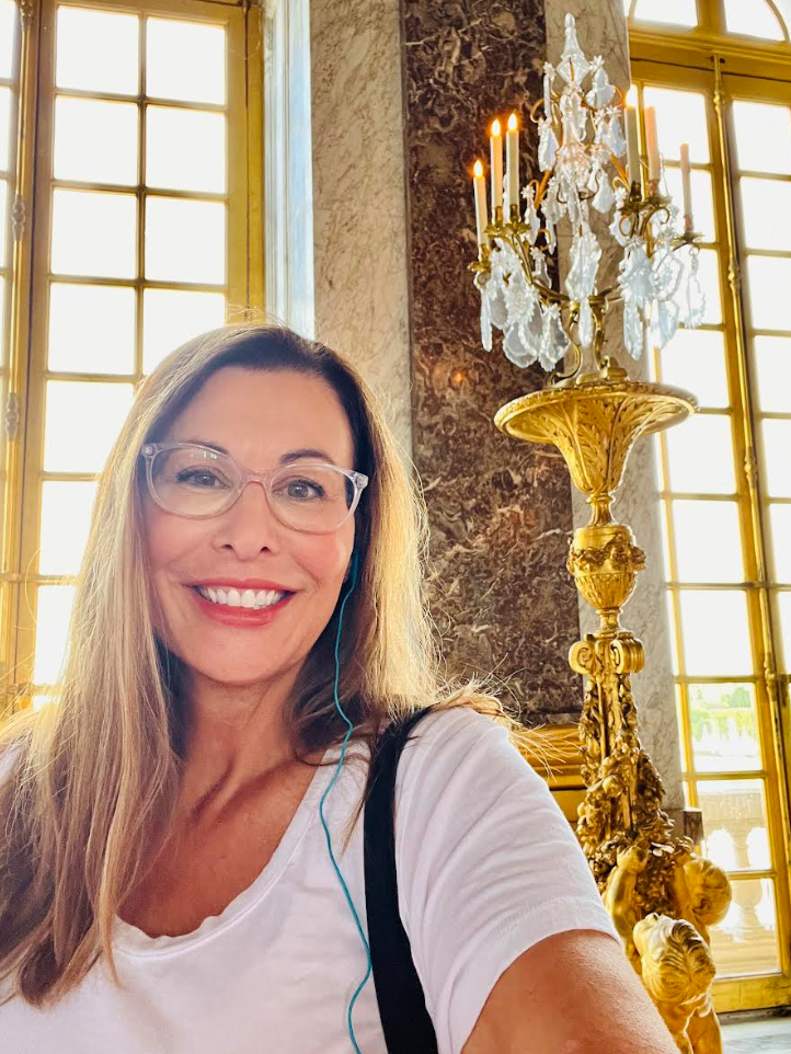 A person smiling while taking a selfie indoors near an ornate chandelier and large windows