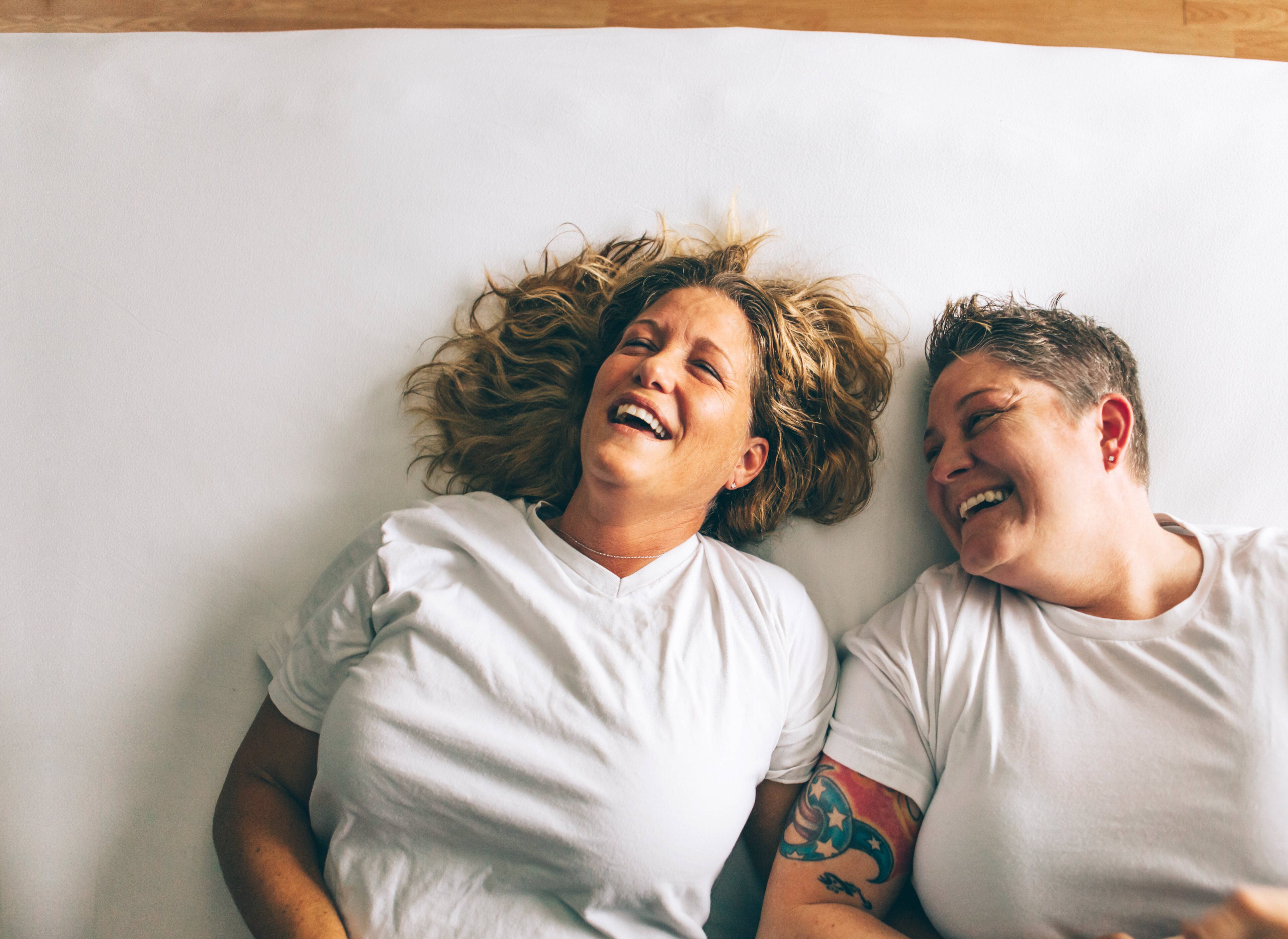 A same-sex couple laying next to each other in bed, laughing
