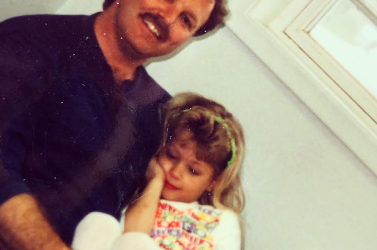 A man with a mustache holds a child sitting on his lap inside a room. The child is resting their head on the man's chest, looking relaxed
