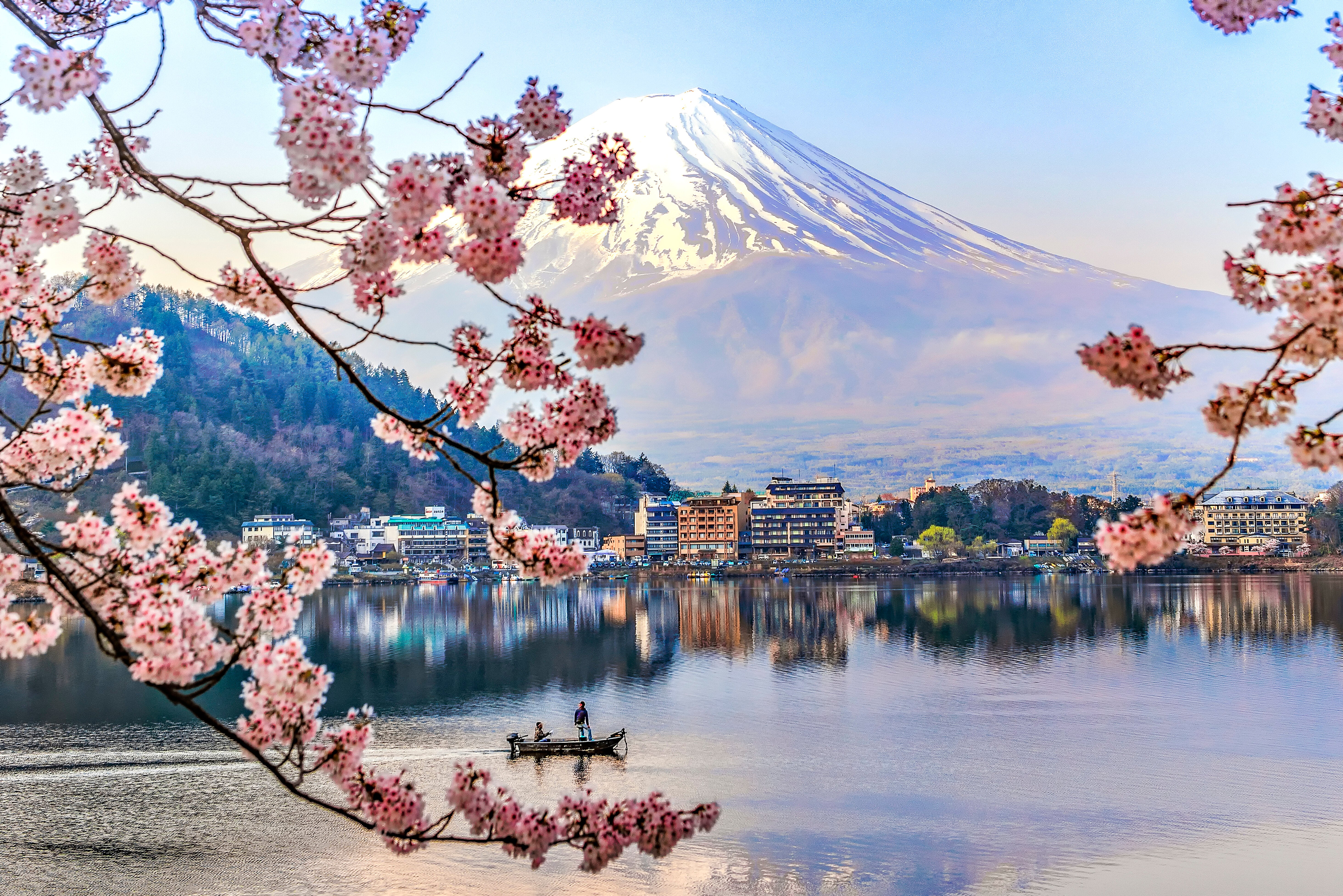 Cherry blossoms frame a serene view of Mount Fuji, reflected in a calm lake, with a small boat and two people in the foreground
