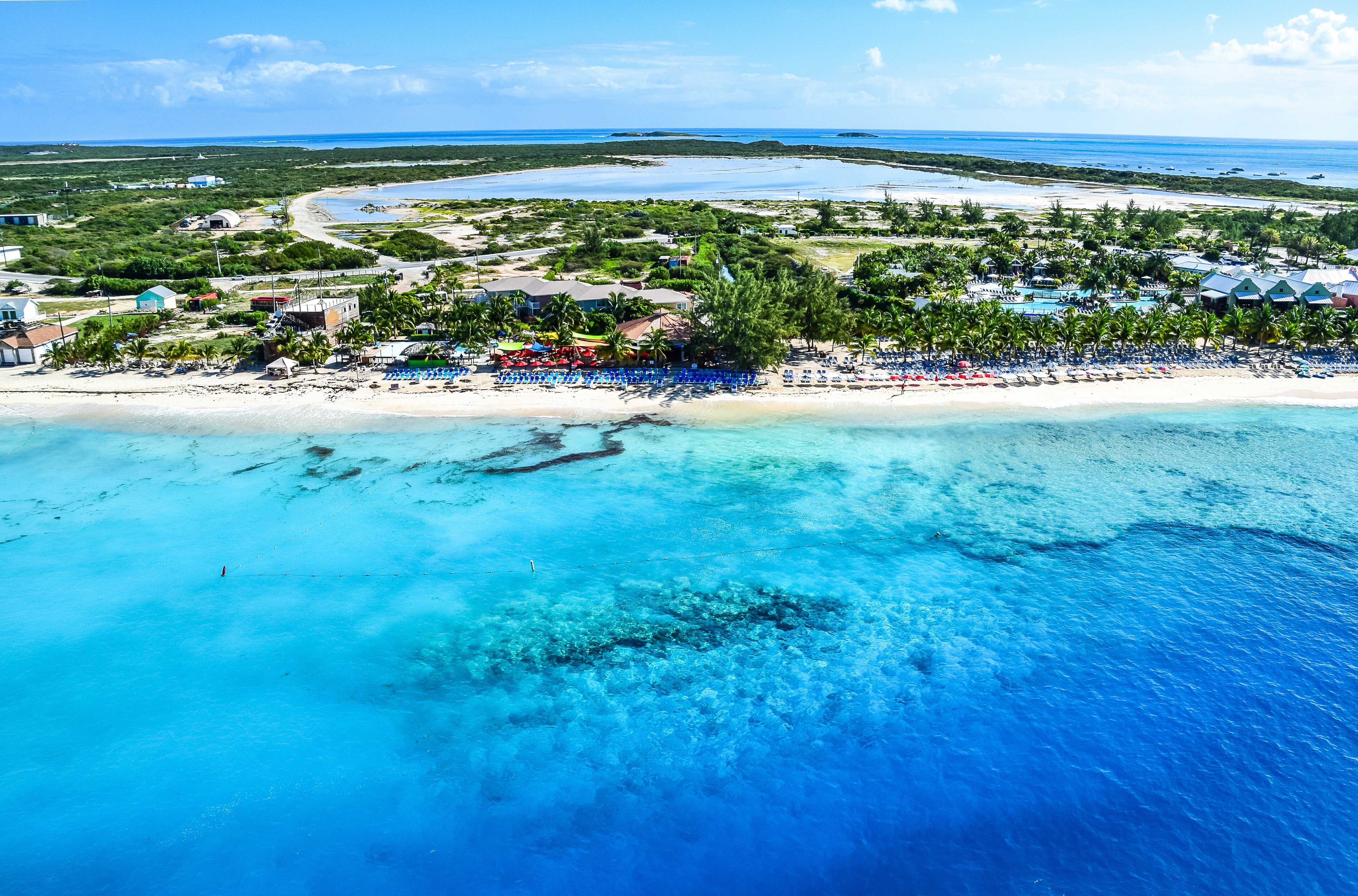 Aerial view of a coastal town with a sandy beach, clear ocean water, and scattered buildings surrounded by greenery
