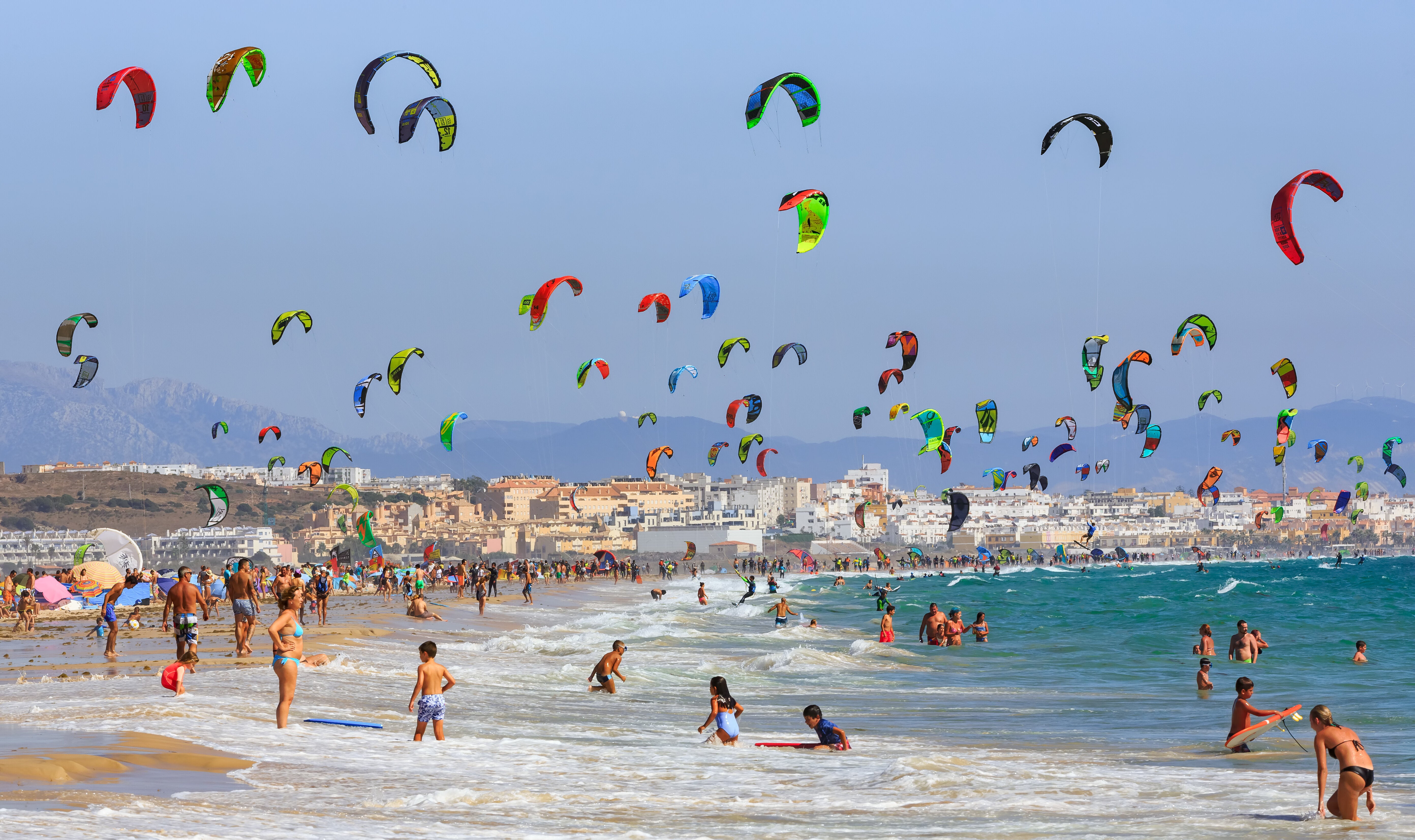 Crowded beach with many people enjoying water and sand; colorful kites fill the sky, suggesting a kite surfing event