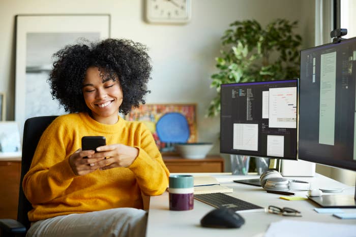 Person at desk with monitors smiling at smartphone