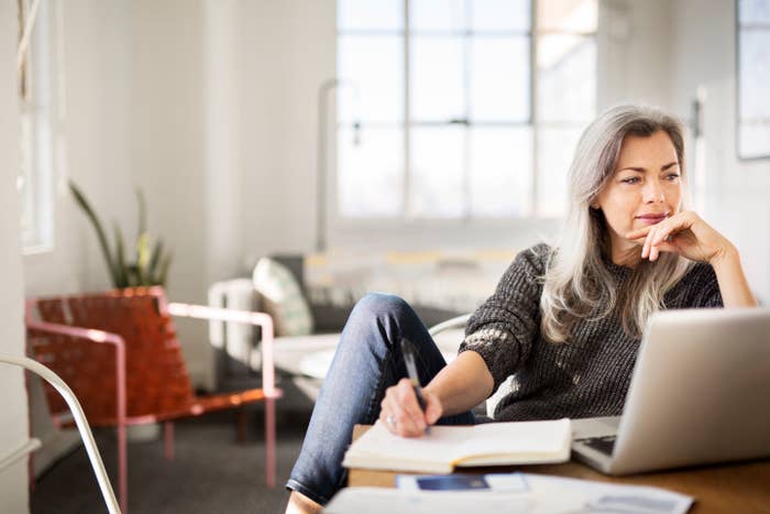 Woman with long gray hair in a cozy living room, sitting at a desk with a notebook, pen, and laptop, appearing thoughtful while looking at the screen