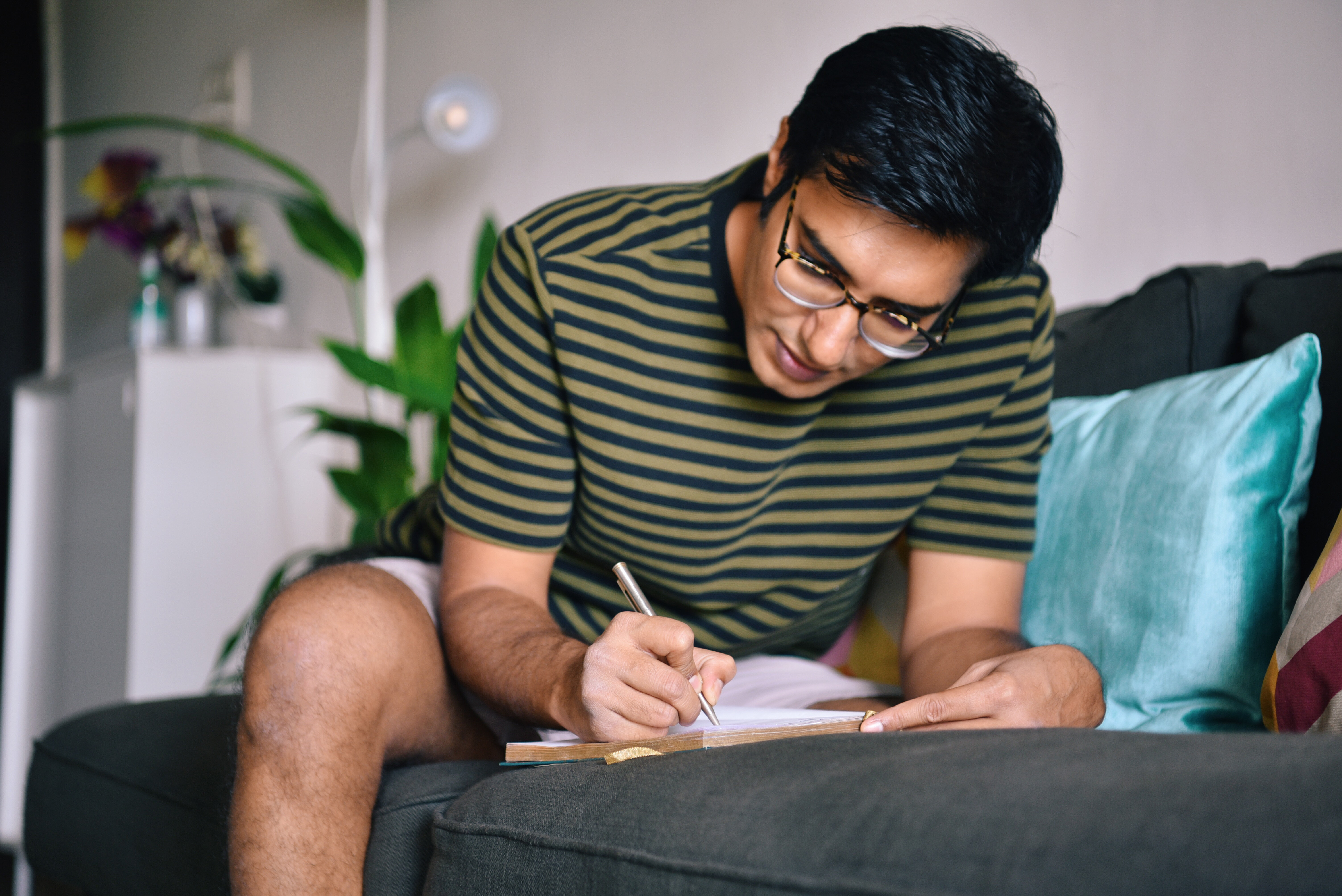 A person wearing glasses and a striped shirt sits on a couch, writing in a notebook, with plants and a lamp in the background