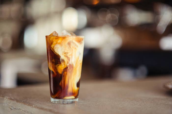 A glass of iced coffee sits on a counter, with swirls of milk mixing into the coffee, creating a marbled effect. The background is blurred