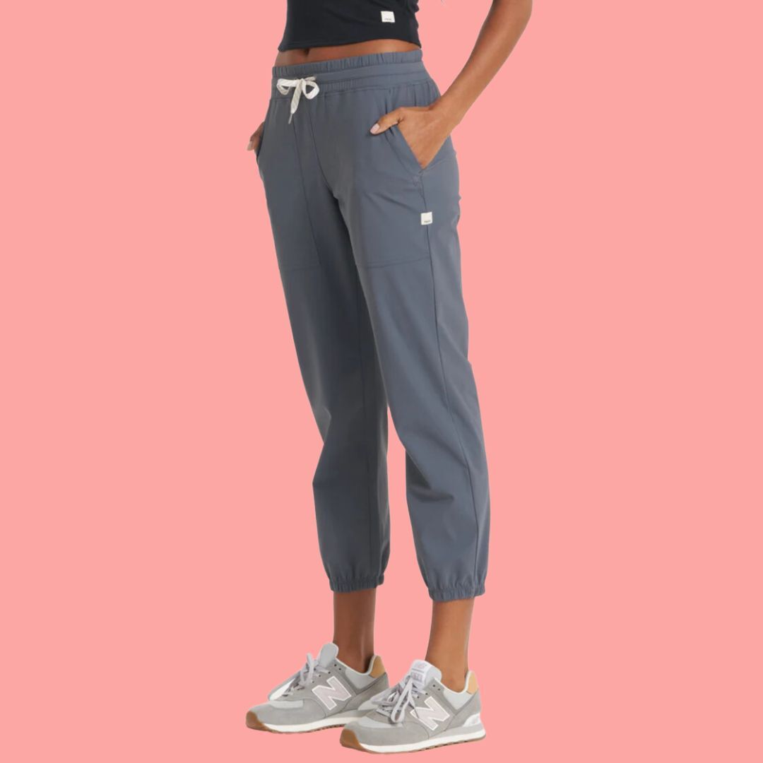 Person wearing casual gray blue trousers and sneakers, standing against a pink background