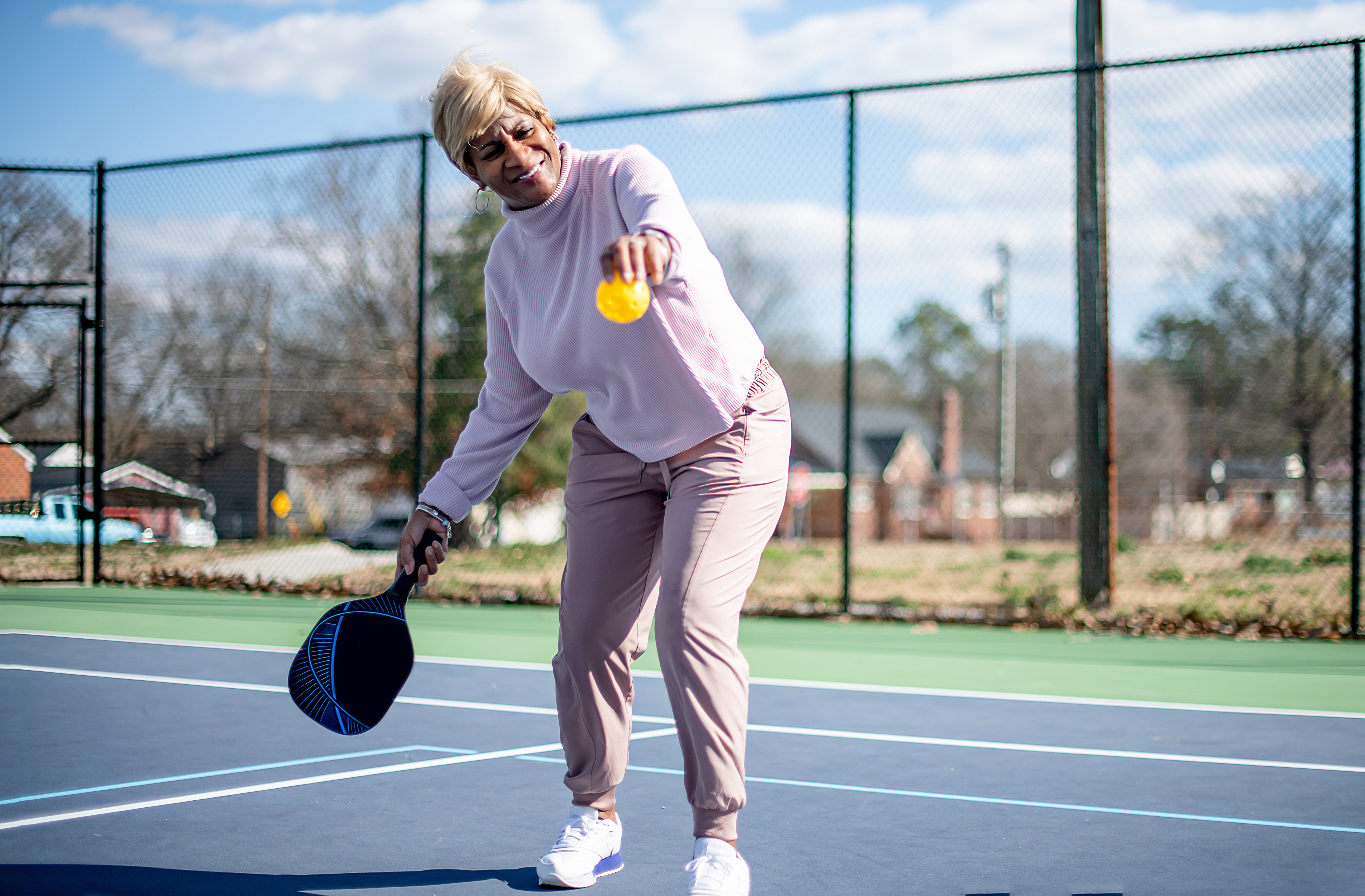 An elderly woman in sporty attire is playing a game of pickleball on an outdoor court, holding a paddle and smiling while reaching for the ball