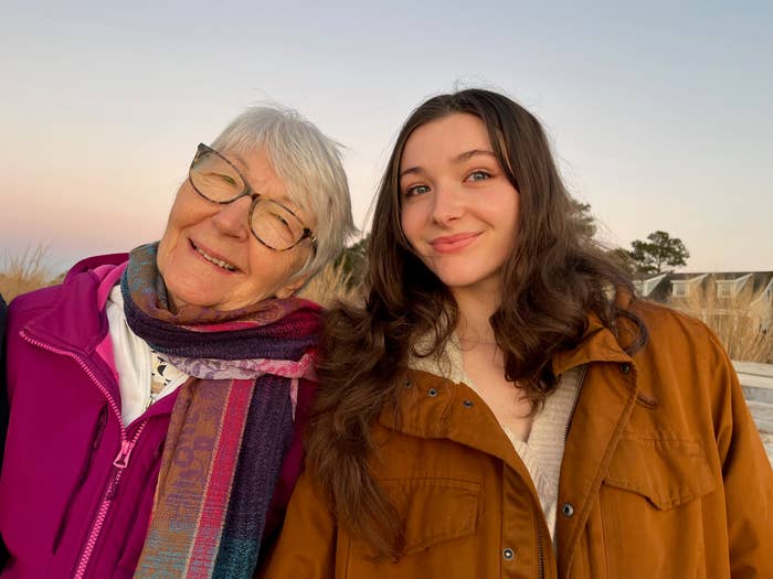 Judy and her granddaughter Emma pose outdoors, both smiling. Judy wears glasses, a scarf, and a jacket, while Emma has loose hair and a casual jacket