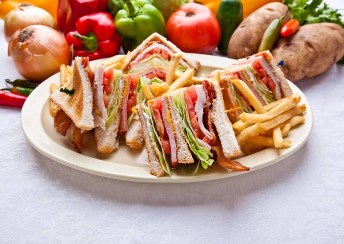 A plate with a club sandwich cut into quarters and a side of french fries, surrounded by fresh vegetables