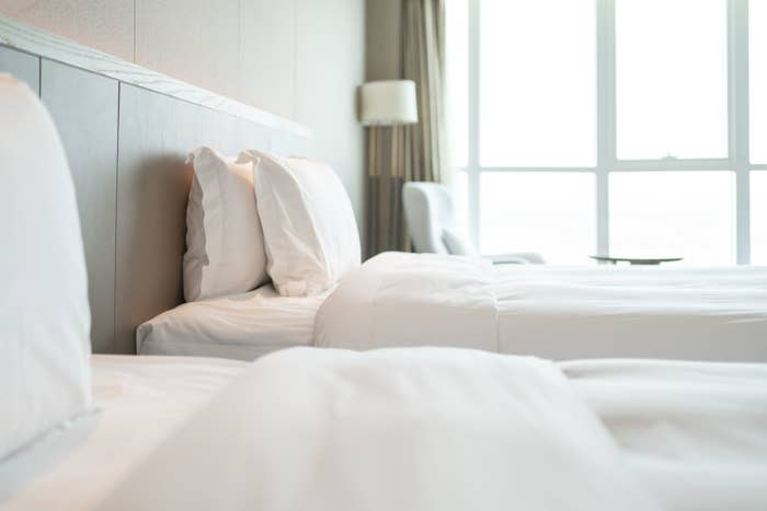 A hotel room with two made beds, pillows neatly arranged, and a chair and lamp by a large window in the background