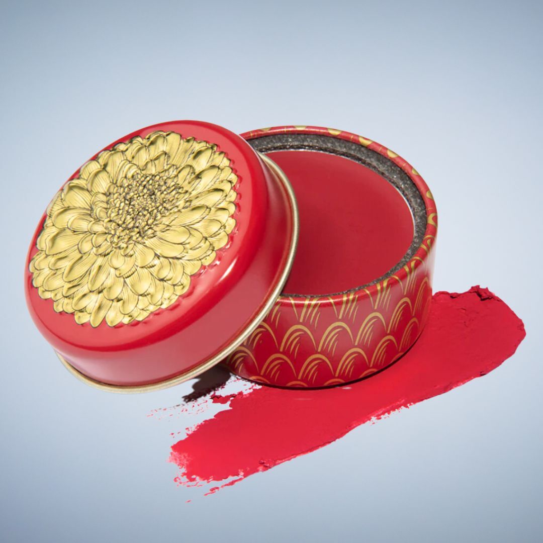Decorative red and gold round tin with embossed lid, open, next to a smear of red cosmetic product