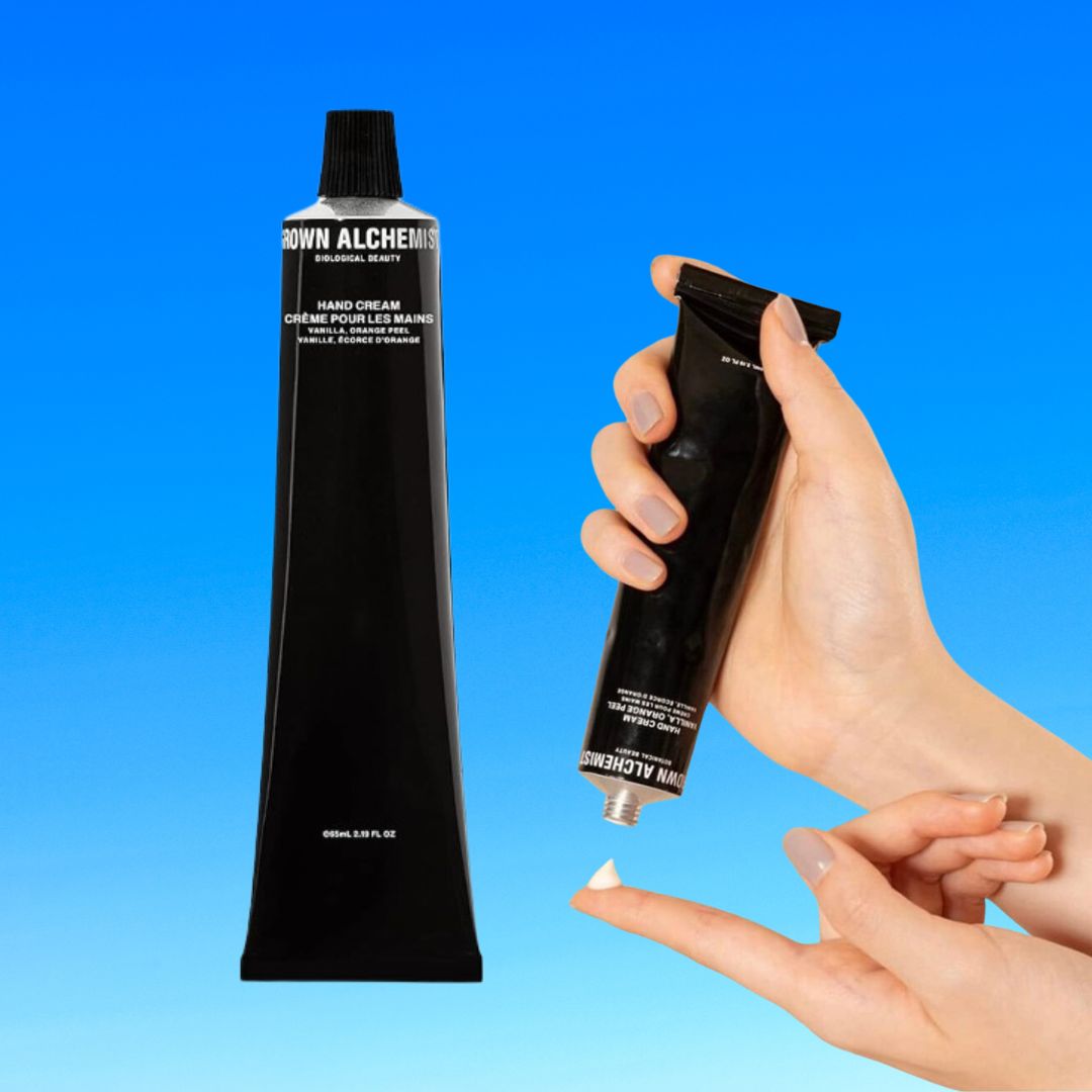 Two hands holding Grown Alchemist hand cream tubes against a blue background. One tube is squeezed with cream on fingertip