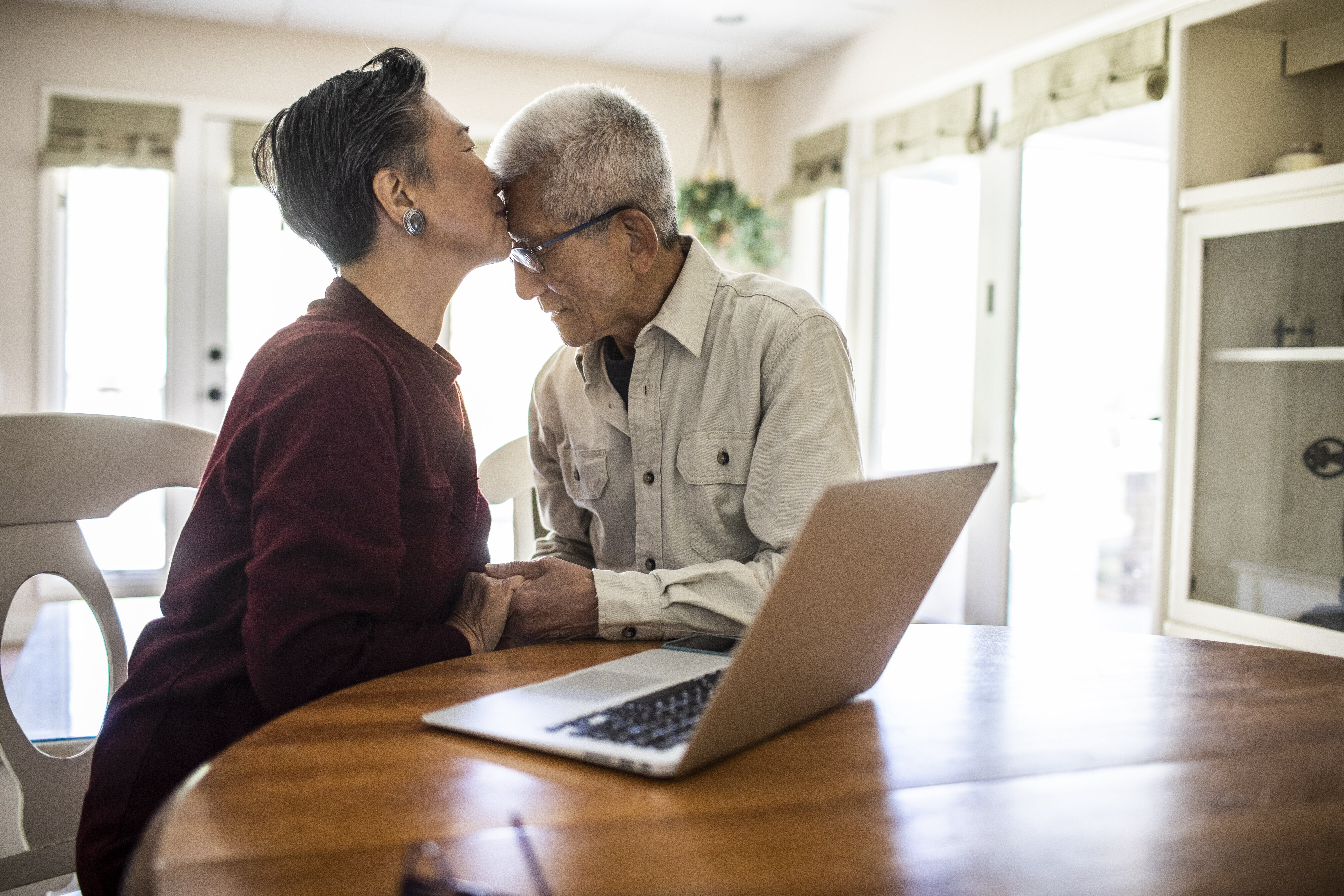 Older couple shares a loving kiss at a table with a laptop, manifesting affection