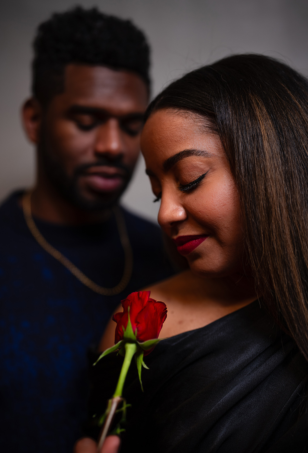 Man behind a woman who holds a rose, both looking down, conveying intimacy and romance