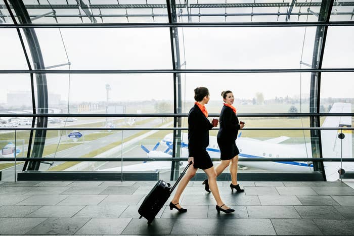 Two flight attendants walking in an airport with an airstrip and planes visible through the glass wall behind them