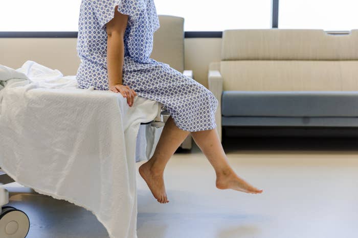 Person in a hospital gown sitting on a bed with legs dangling off the side, in a medical setting