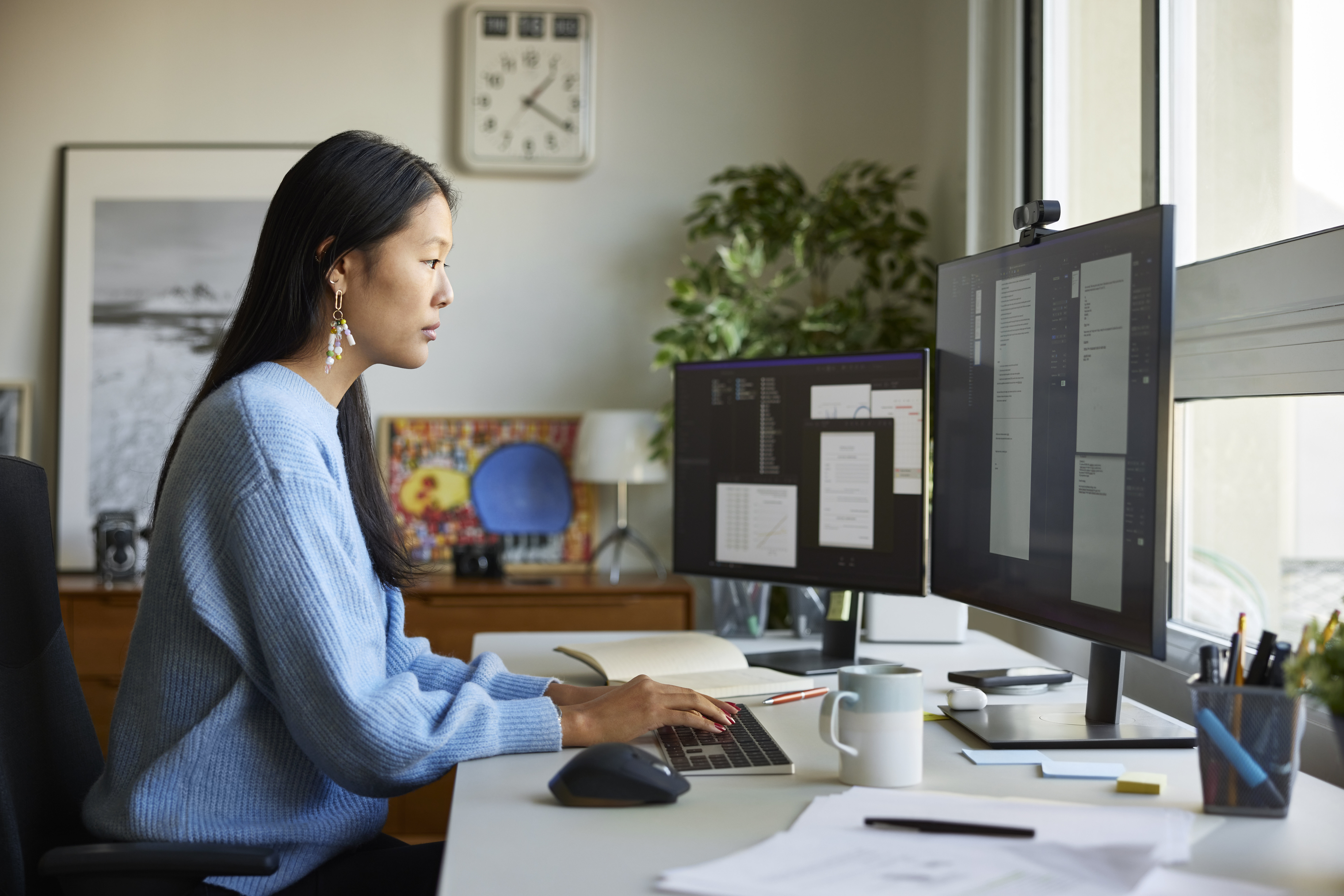 Woman at desk with dual monitors working on a document. She&#x27;s focused, wearing a casual sweater