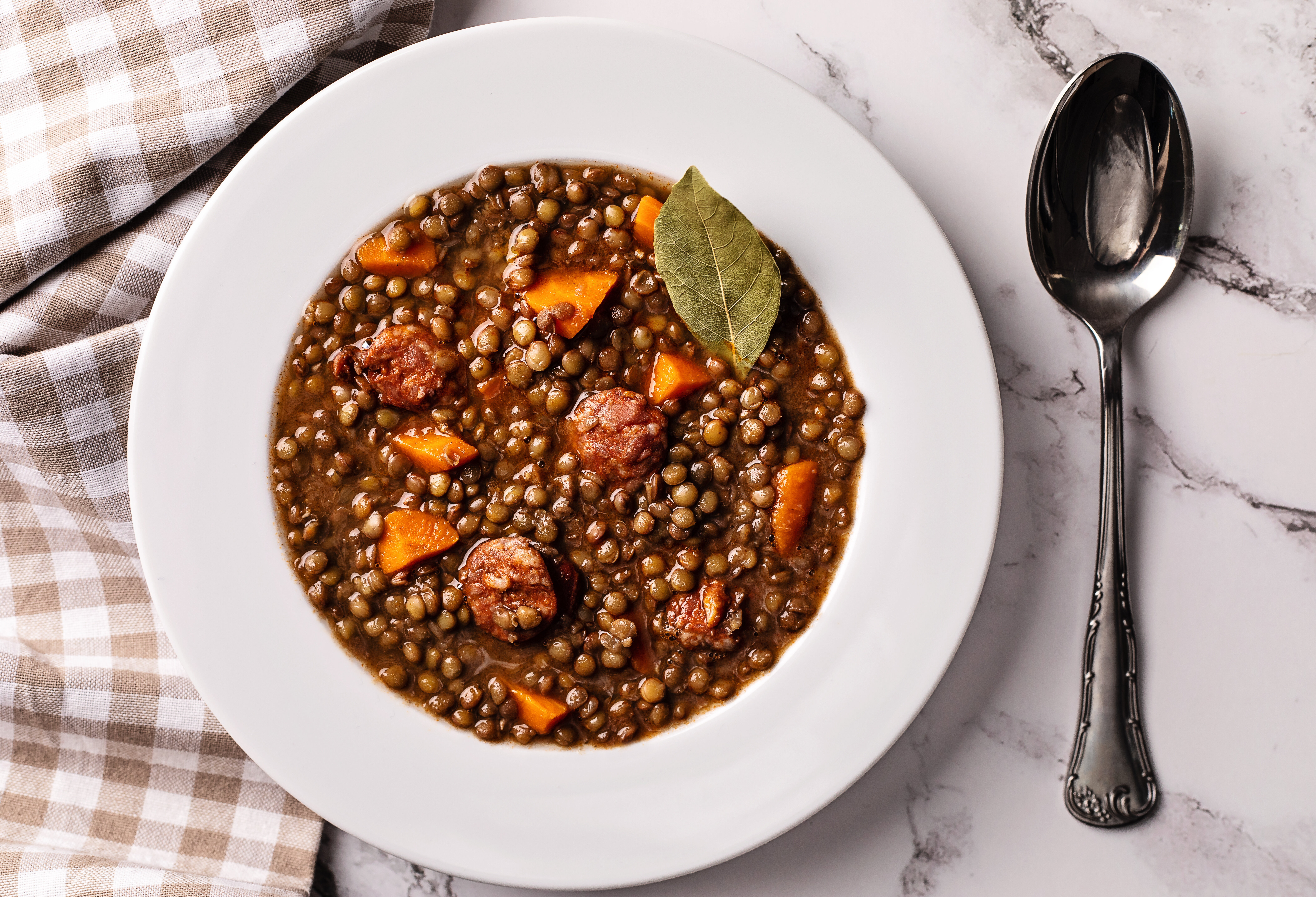 A bowl of lentil stew with chunks of sausage and carrot, served on a white plate with a spoon on the side