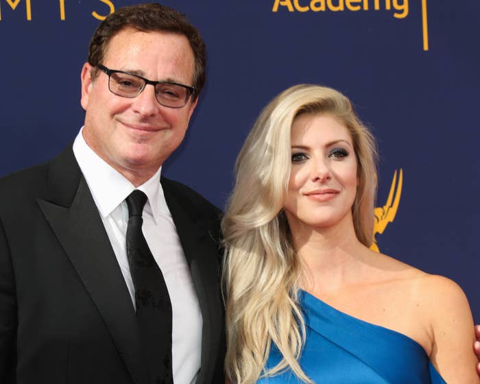 Bob Saget in a black suit and tie stands next to Kelly Rizzo in a one-shoulder dress at an event