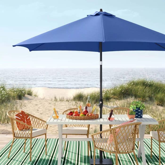 A green striped outdoor rug, styled with a white table, rattan chairs, and a blue umbrella, on a beach looking out over the water