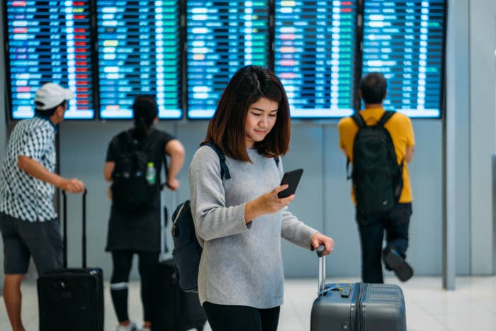 Woman checking her phone while standing with luggage at airport departure board