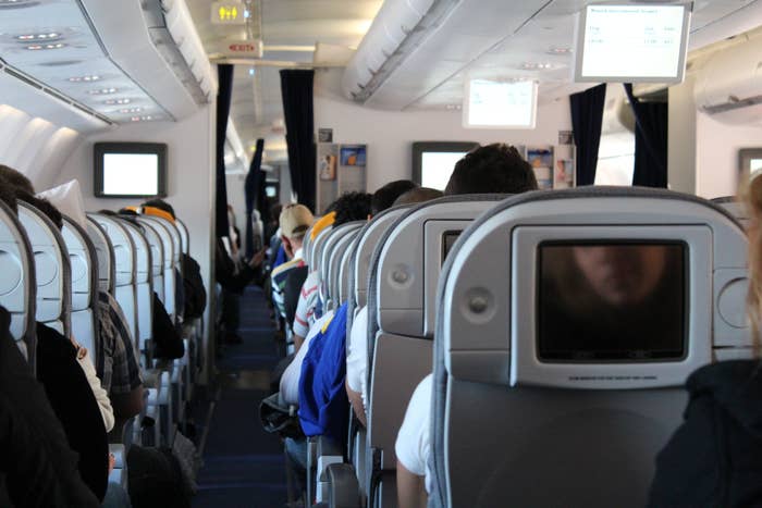 Passengers seated inside an aircraft cabin with overhead screens and aisle visible