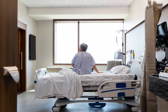Person in hospital gown sitting on a bed facing the window in a medical room