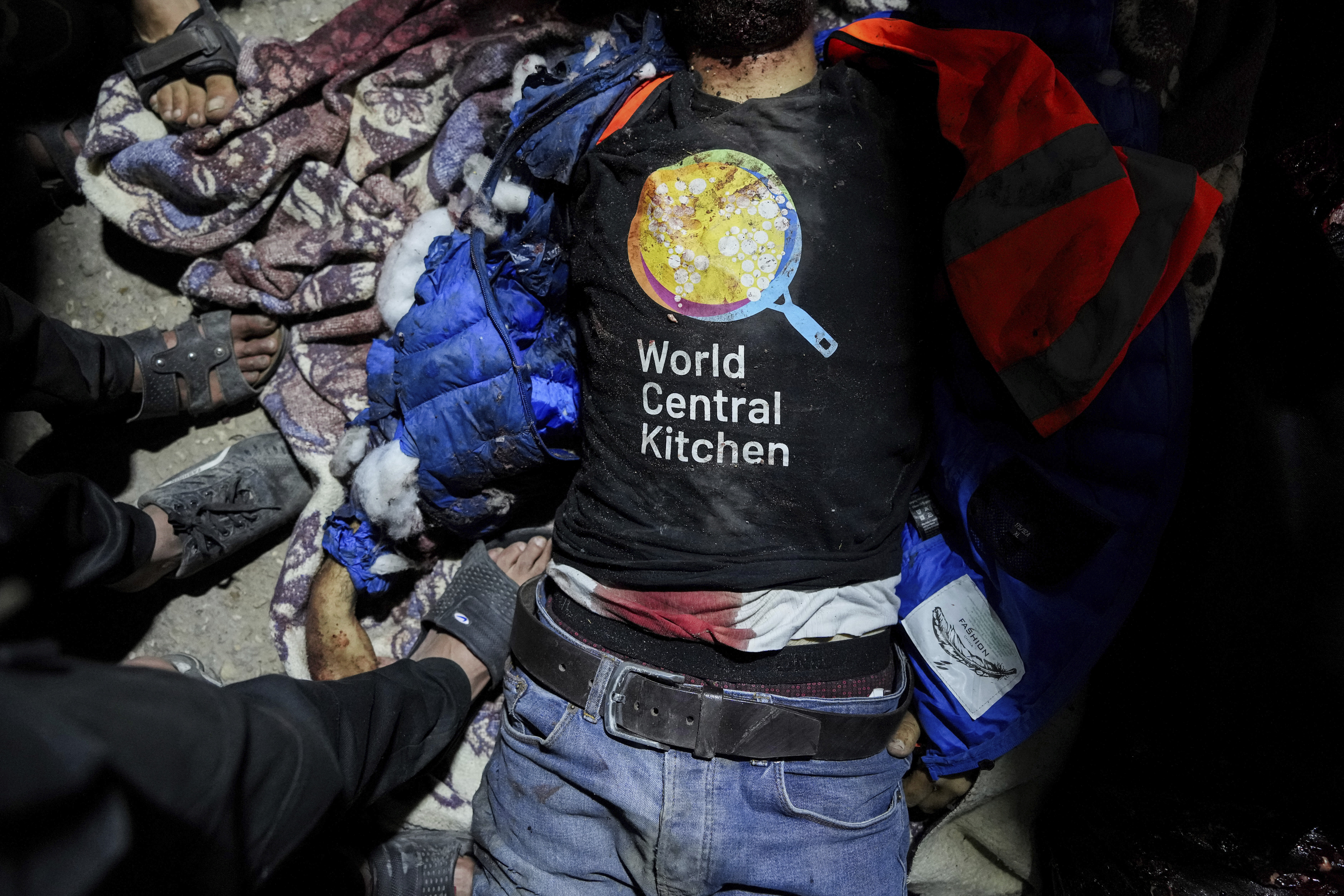 Person lying on ground with &quot;World Central Kitchen&quot; shirt, surrounded by helping hands, debris visible