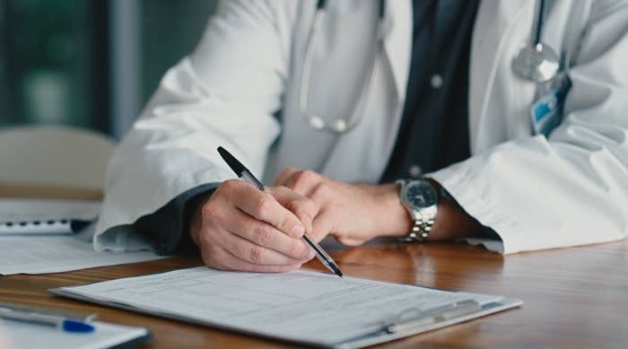 A doctor in a white coat is writing on a clipboard at a wooden desk, with a stethoscope around their neck
