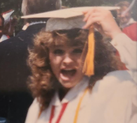 Graduate in cap and gown, smiling and raising the cap tassel, surrounded by a crowd in a celebratory setting