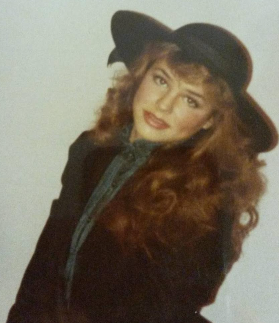 Person with long hair wearing a wide-brimmed hat and stylish jacket, posing with a tilted head and soft expression