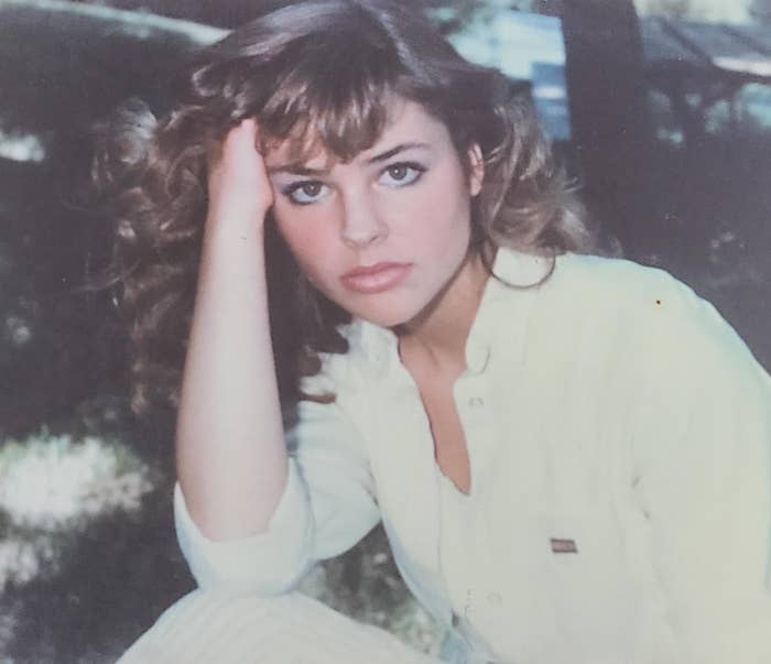 Person with wavy hair poses outdoors, looking at the camera with a hand rested on their head, wearing a casual button-up shirt