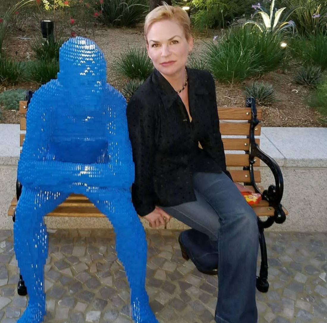 A woman sits on a bench beside a blue abstract sculpture of a person, set in a public outdoor space