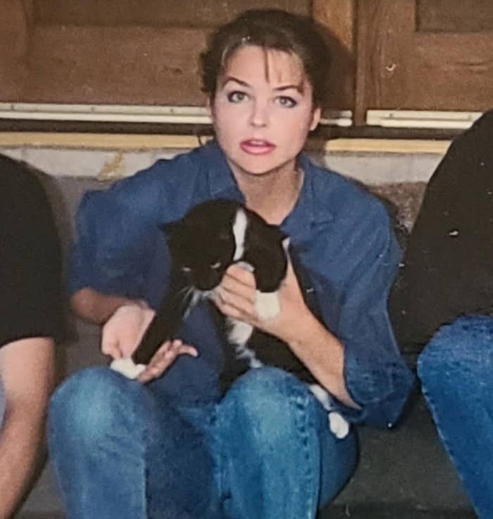 Woman in casual denim holding a black-and-white cat, sitting indoors with others partially visible