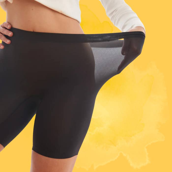 Person wearing stretchy achromatic  shapewear shorts, demonstrating flexibility by pulling the cloth  distant  from the waist, against a yellowish  background