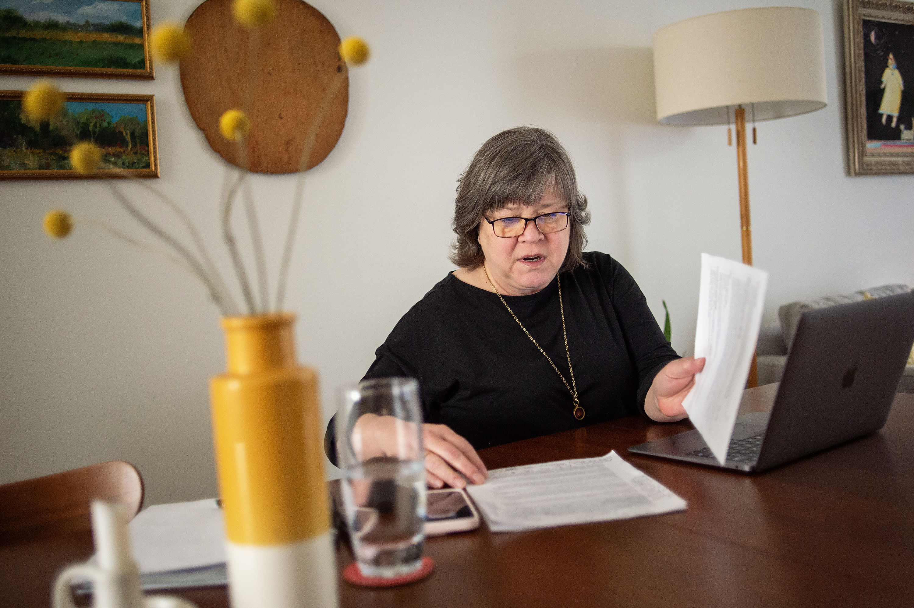 An older woman reads documents at a desk with a laptop, glass of water, and vase with yellow flowers. She is focused and appears to be working