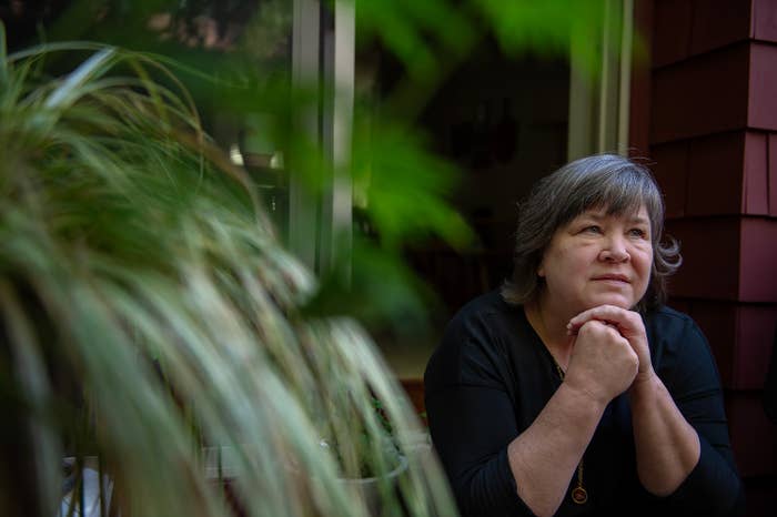 A woman with gray hair, wearing a black top, sits with her chin resting on her hands, looking thoughtfully into the distance by a window