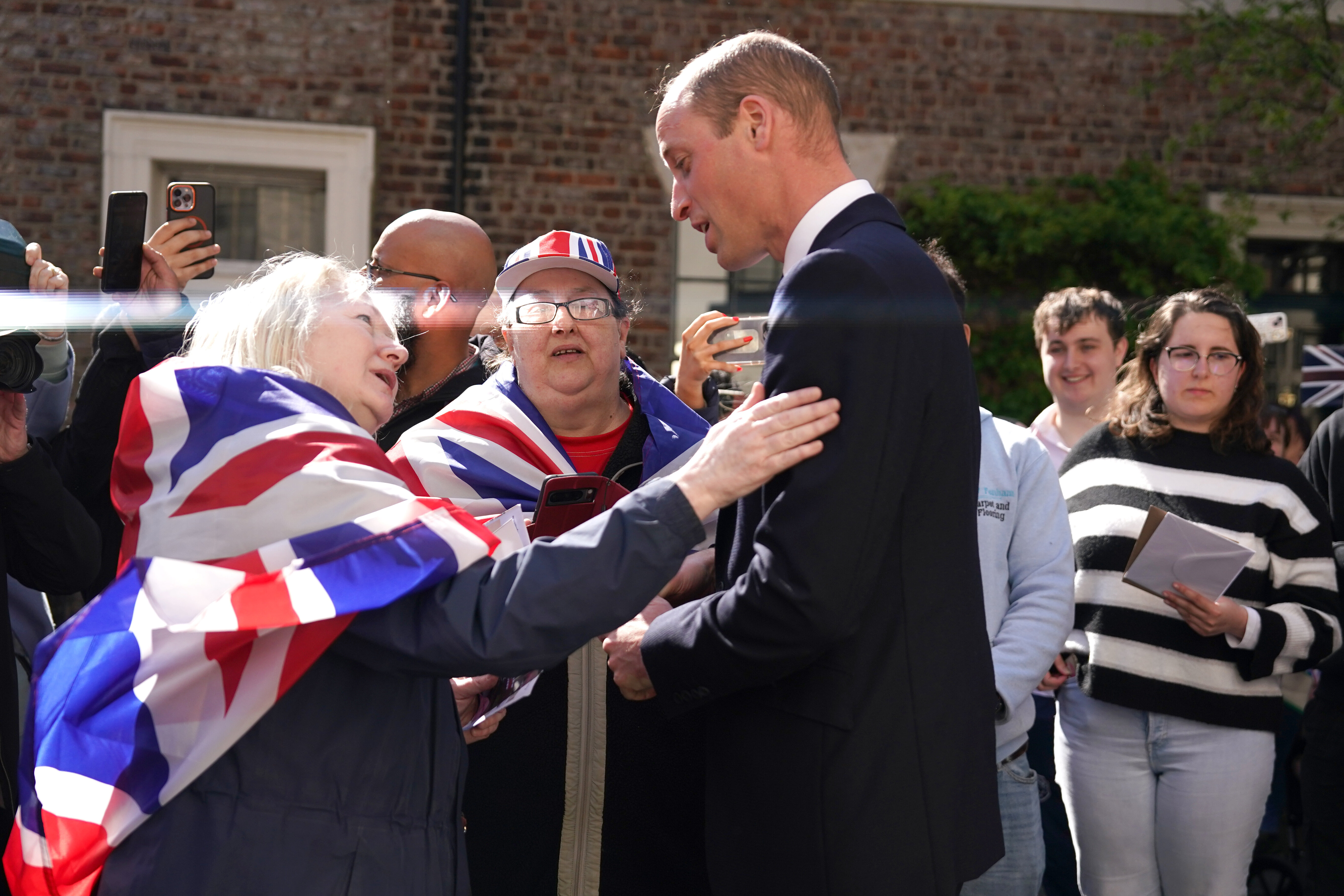 Prince William engages with a cheering crowd, people taking photos; a woman in a Union Jack scarf directly interacts with him