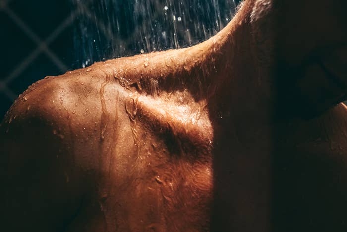 A close-up of a person's upper body and neck under running water in the shower, capturing the wet skin and droplets. The person's face is partially visible