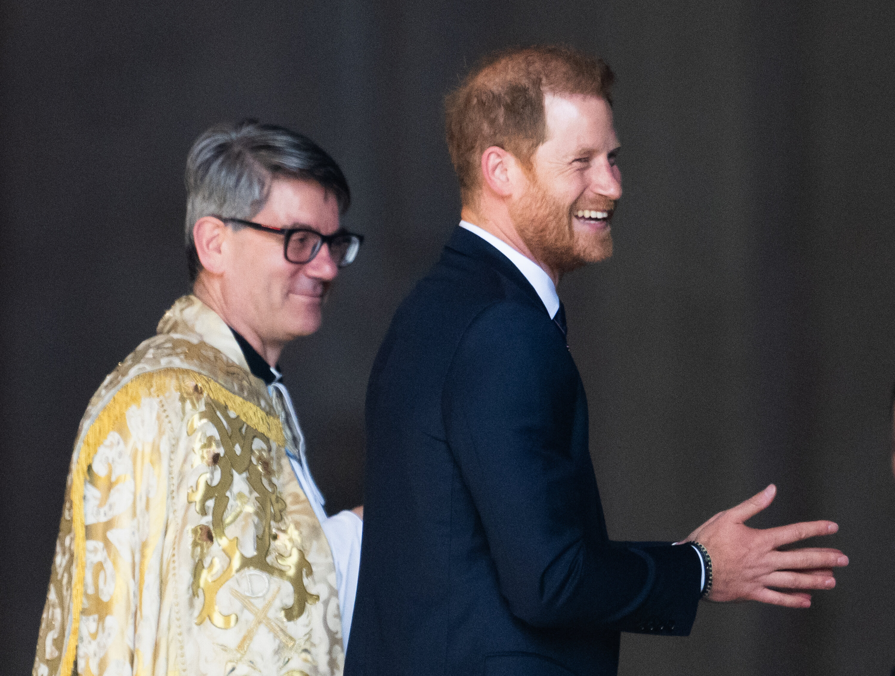 Prince Harry in a dark suit smiling and walking beside a person in ceremonial robes