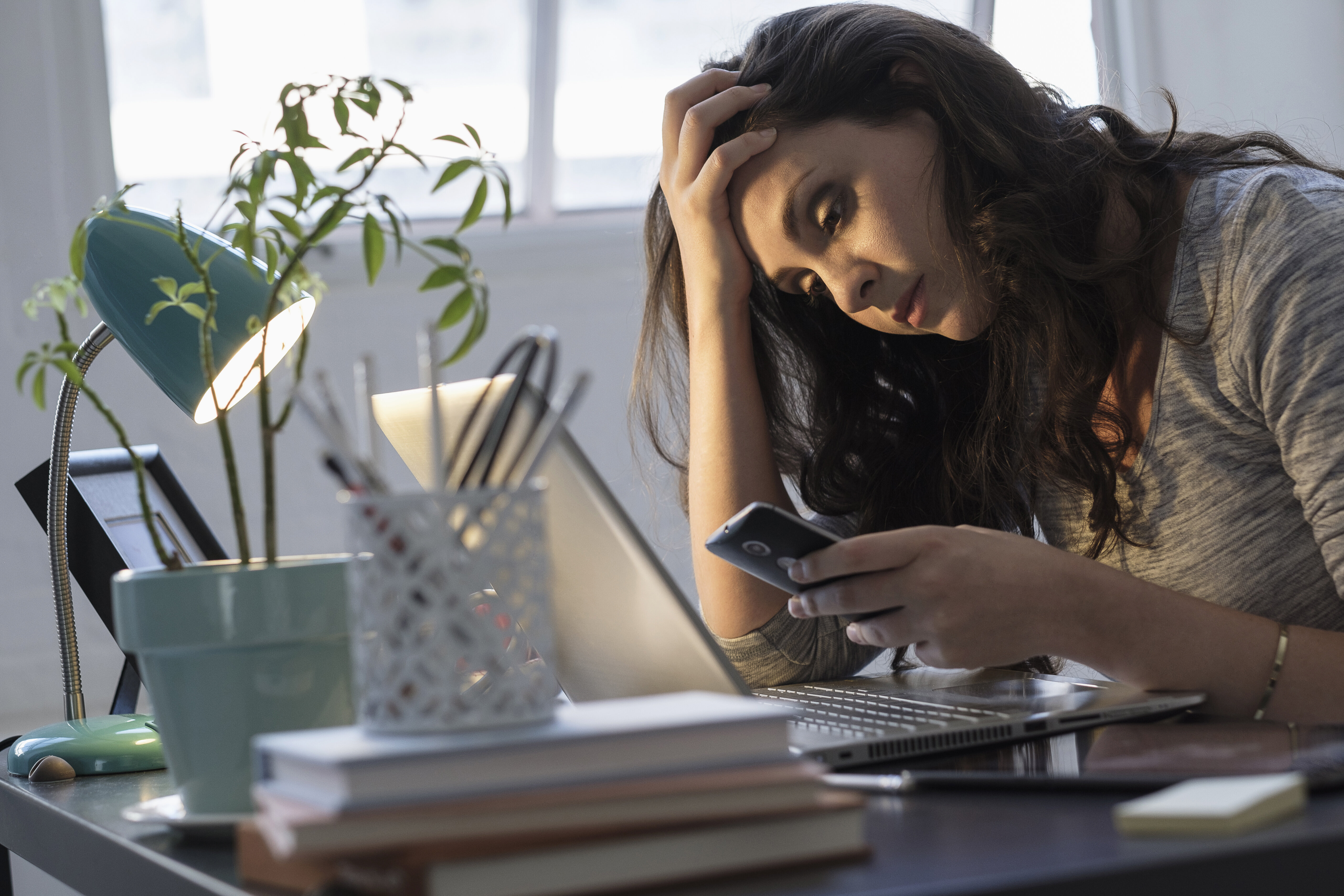 Woman in a gray shirt looks stressed while holding a phone and sitting at a desk with plans, books, a plant, and a lamp