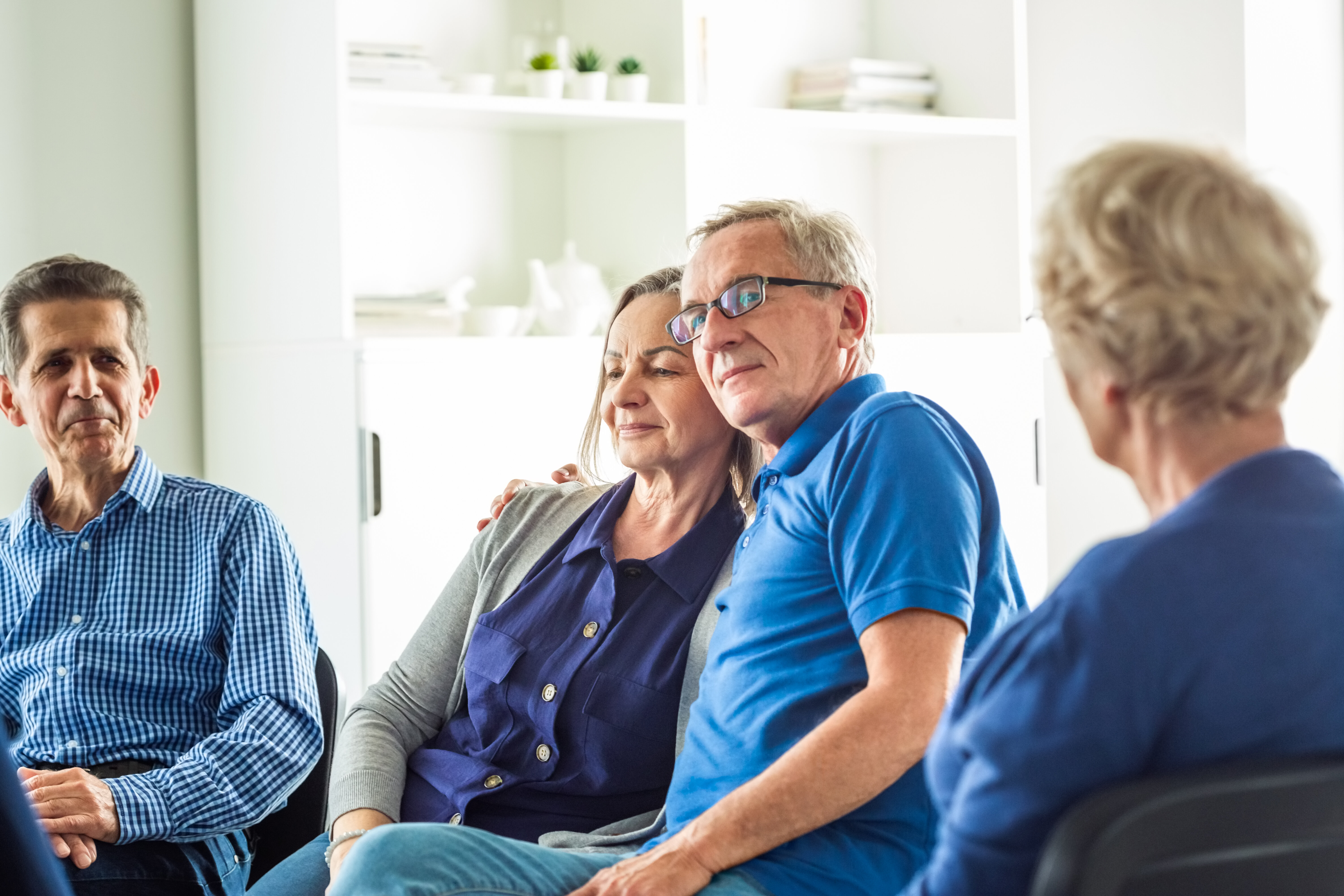 People sit in a support group. A man in glasses and a blue shirt embraces a woman beside him, both looking content