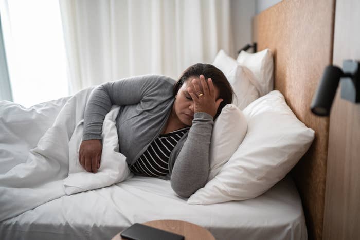 A person is lying in bed with their hand on their forehead, appearing to be in pain or discomfort. A wooden headboard and a side table are visible