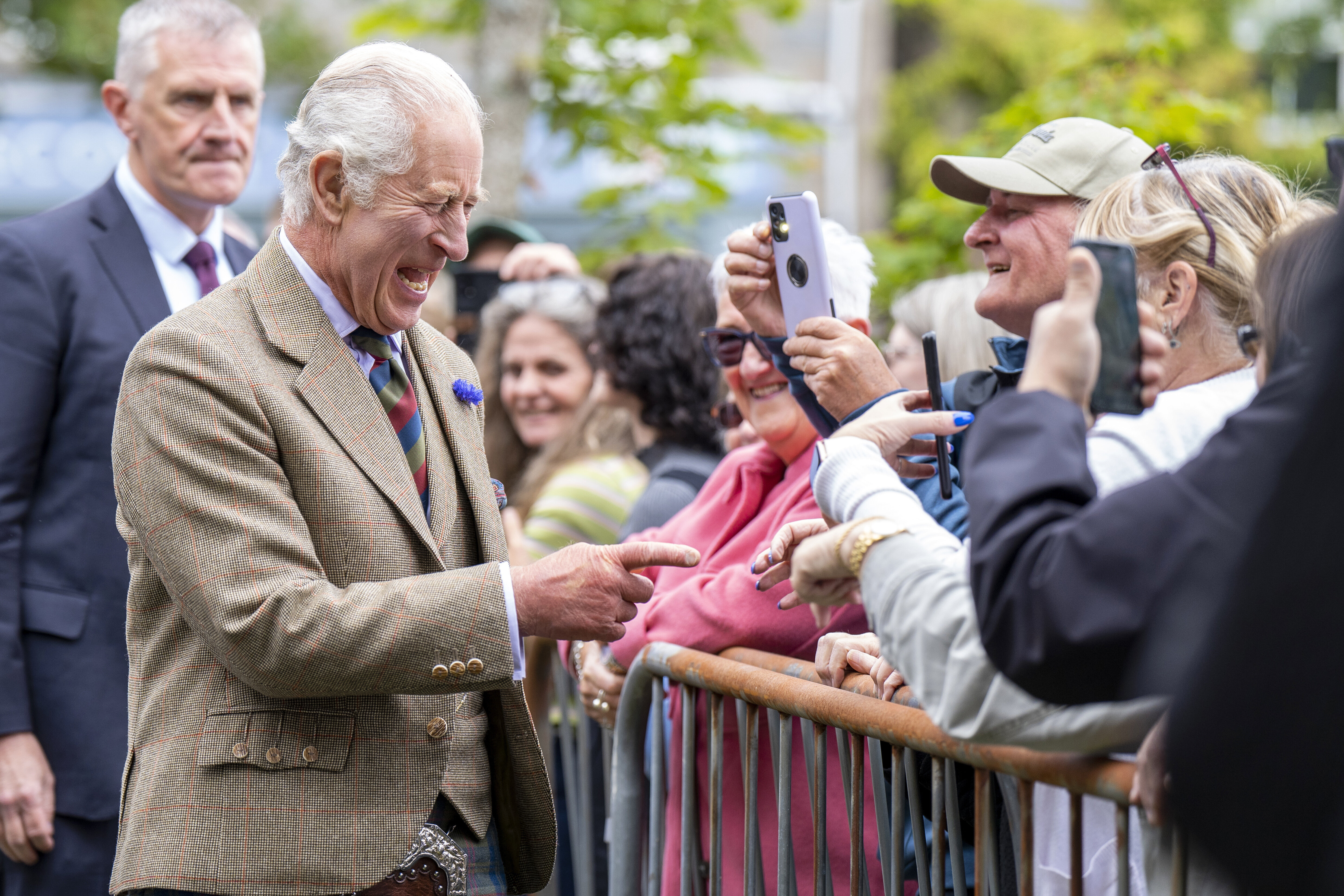 King Charles III smiling and chatting with fans behind a barricade, as an older man stands guard nearby. King Charles III wears a patterned suit