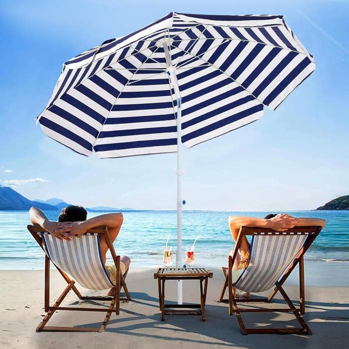 Two people relax on lounge chairs under a striped beach umbrella, facing the ocean with drinks on a small table between them