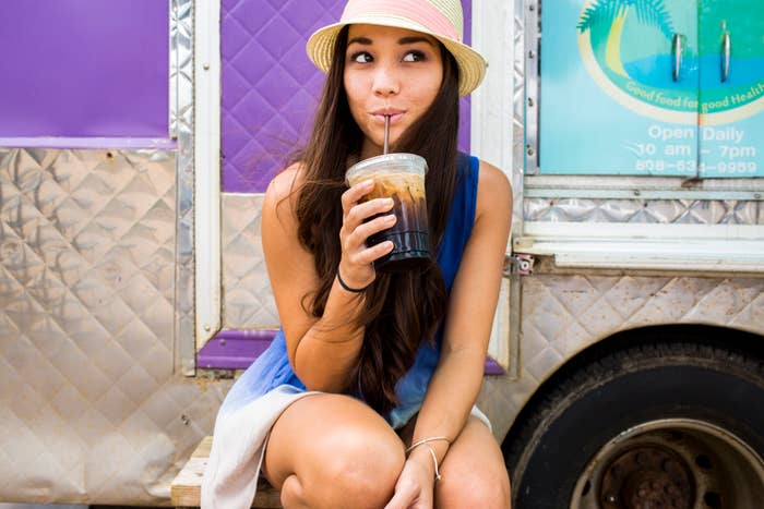 A woman in casual summer attire, straw hat, sipping iced coffee near a food truck. The truck sign reads, &quot;Good food for good health. Open daily 9:30 am - 7 pm.&quot;