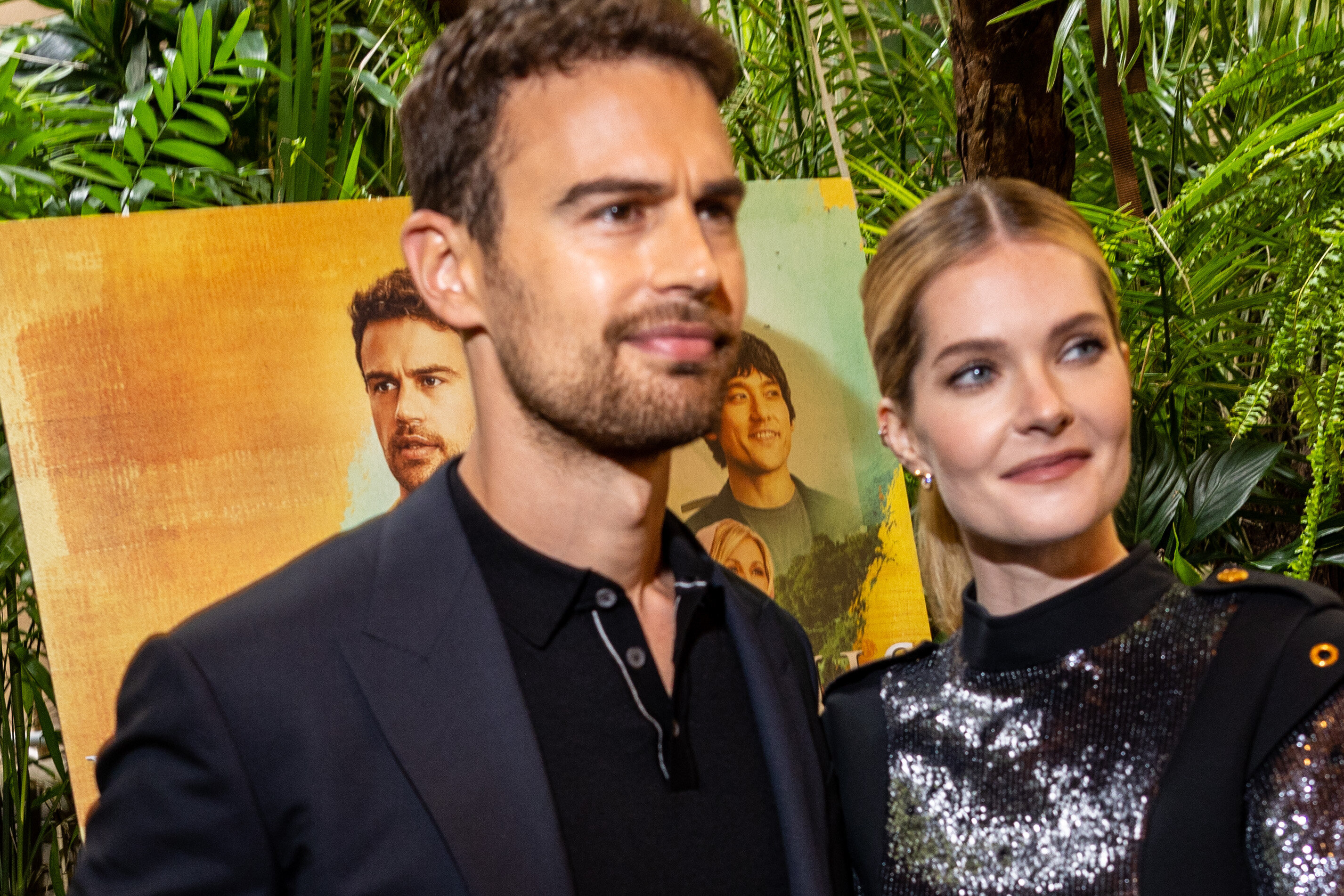 Theo James in a black suit and Meghann Fahy in a sequined dress at a red carpet event with a colorful promotional poster in the background