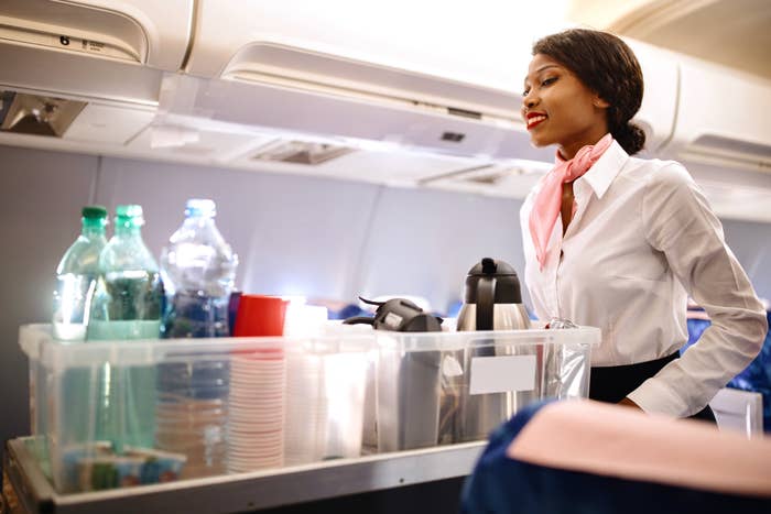 A flight attendant with a pink scarf serves drinks and refreshments from a trolley on an airplane